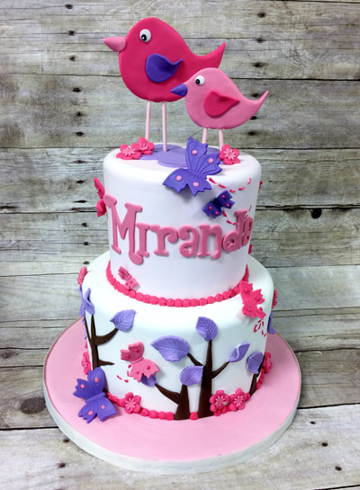 2 tier baby shower cake for a girl. Features 2 birds on top and cake is decorated with trees in colors of pink and purple.