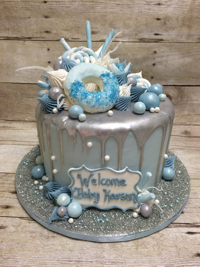 silver and blue drip icing baby shower cake with blue icing doughnut and lots of decorations on top.