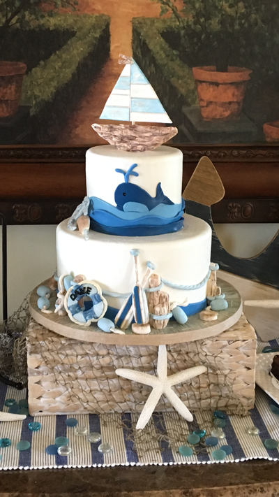 2 tier baby shower cake with nautical theme. Sail boat on top, whale in water scene on top tier and surrounding bottom tier are row boat paddles, buoys, lifesaver buoy, and nautical rope.