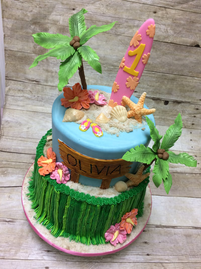 2 tier birthday cake with an Hawaiian theme. bottom tier looks like a grass skirt, top layer has a chocolate surfboard, sea shells and palm trees. 