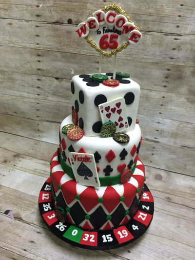 Casino theme 3 tiered birthday cake. bottom layer is surrounded by roulette wheel number slots in red black and green. 2nd tier has hearts and spade playing card, top tier is cake in a cube shape to look like a dice and topped with a chocolate Las Vegas sign.