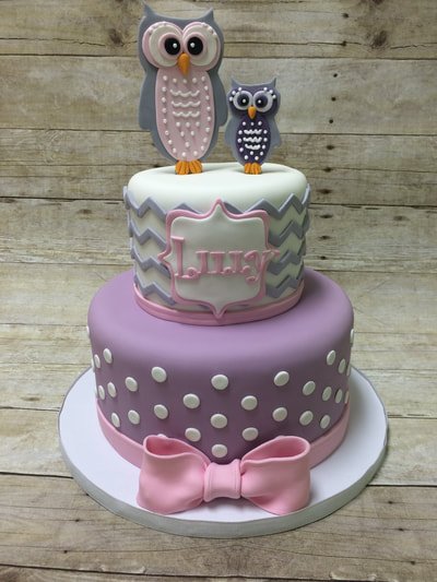 owl theme 2 tier baby shower cake. top tier has white background on cake with wavy gray lines. Bottom tier is purple background with white polka dots all finished off with a pink bow.