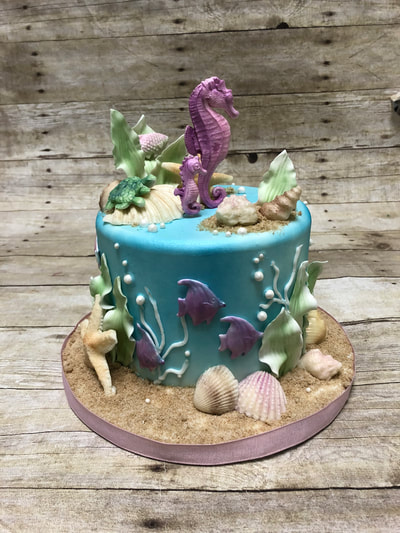 under the sea baby shower cake. blue fondant covered cake looks like the water for a sea horse on top and kelp plants, fondant fish and plants on the side of the cake and chocolate sea shells around the base.