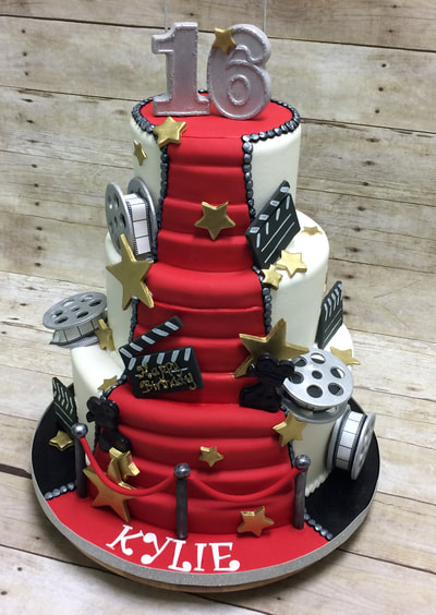 3 tier birthday cake with red carpet stairs from top to bottom and movie props all around including red velvet ropes.